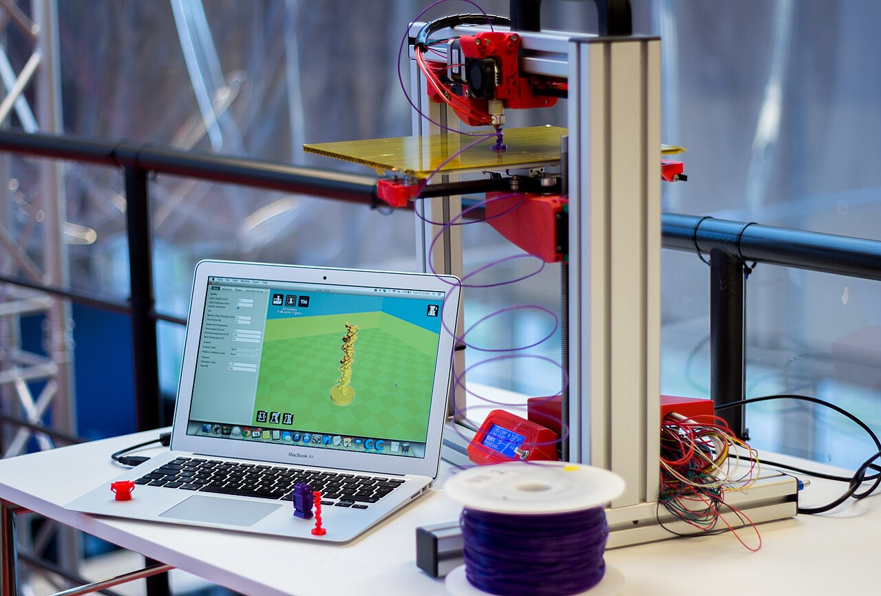 Ноутбук с 3D-моделью на экране рядом с работающим 3D-принтером и катушкой пластика